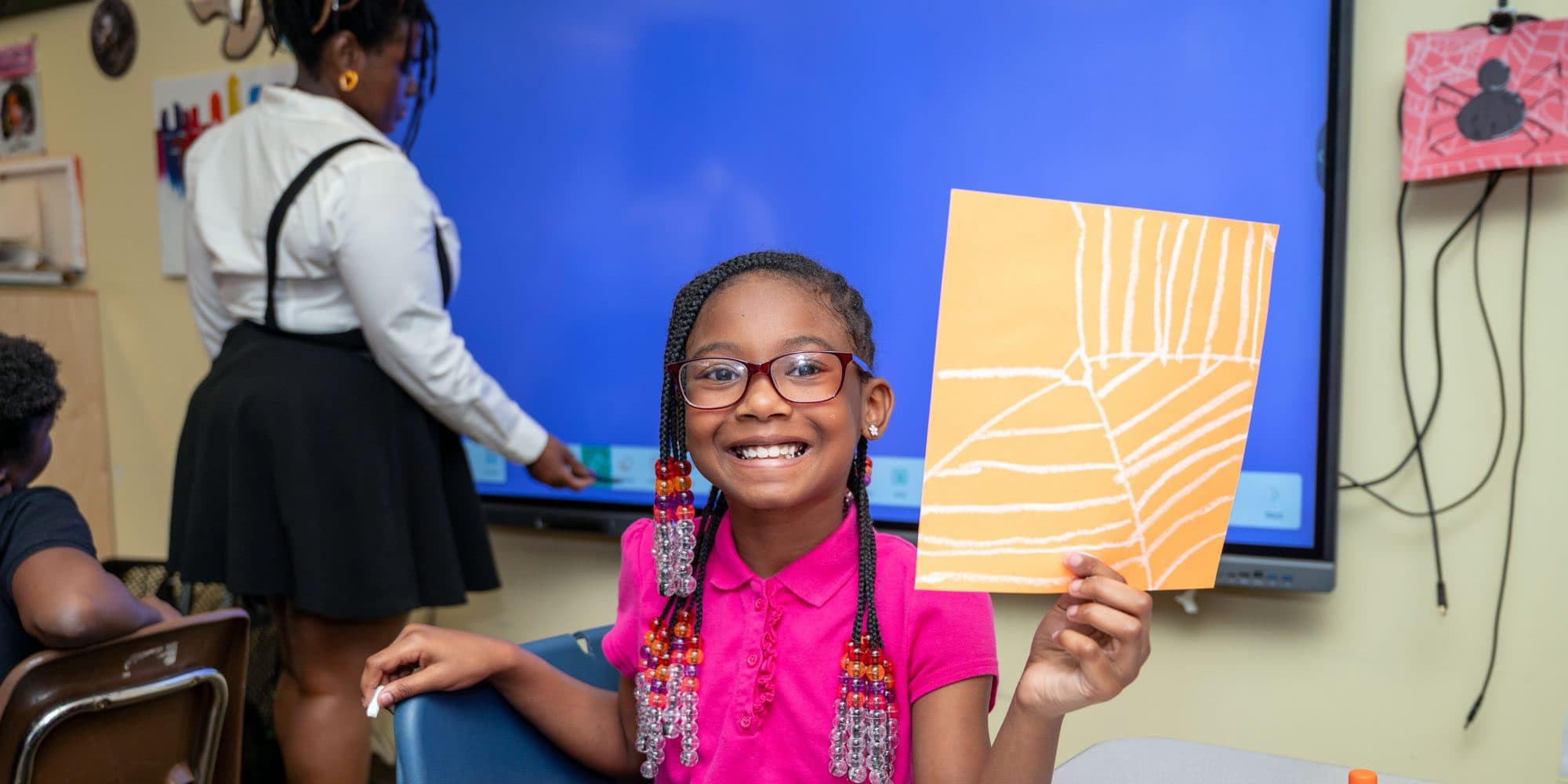 Student shows artwork while a teacher works at the board behind them.