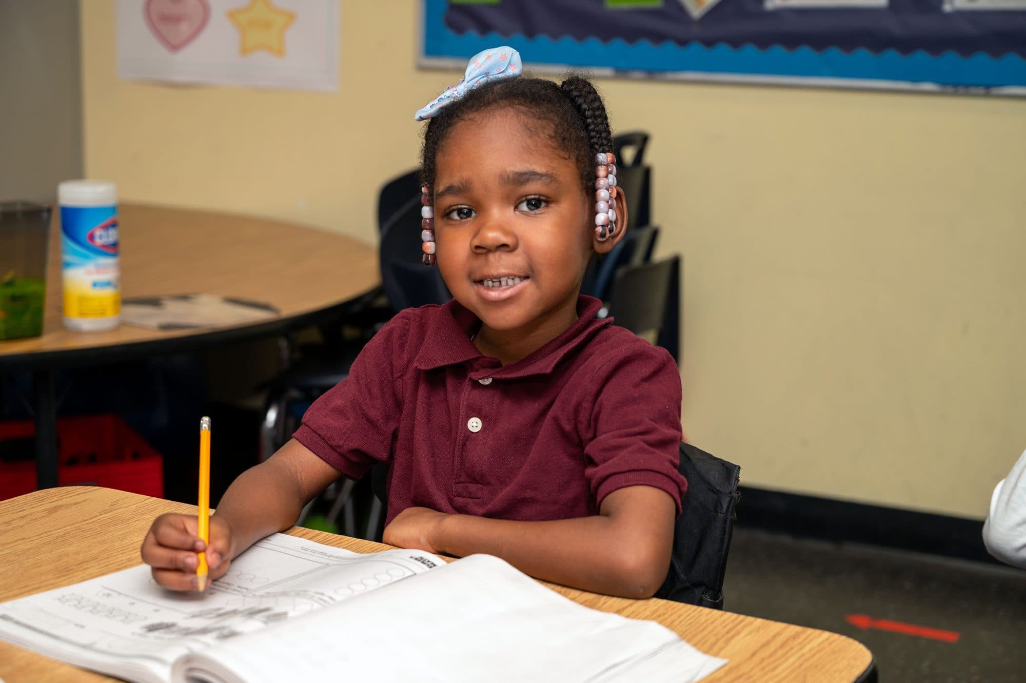 Young student holds a pencil over a workbook at a classroom desk.