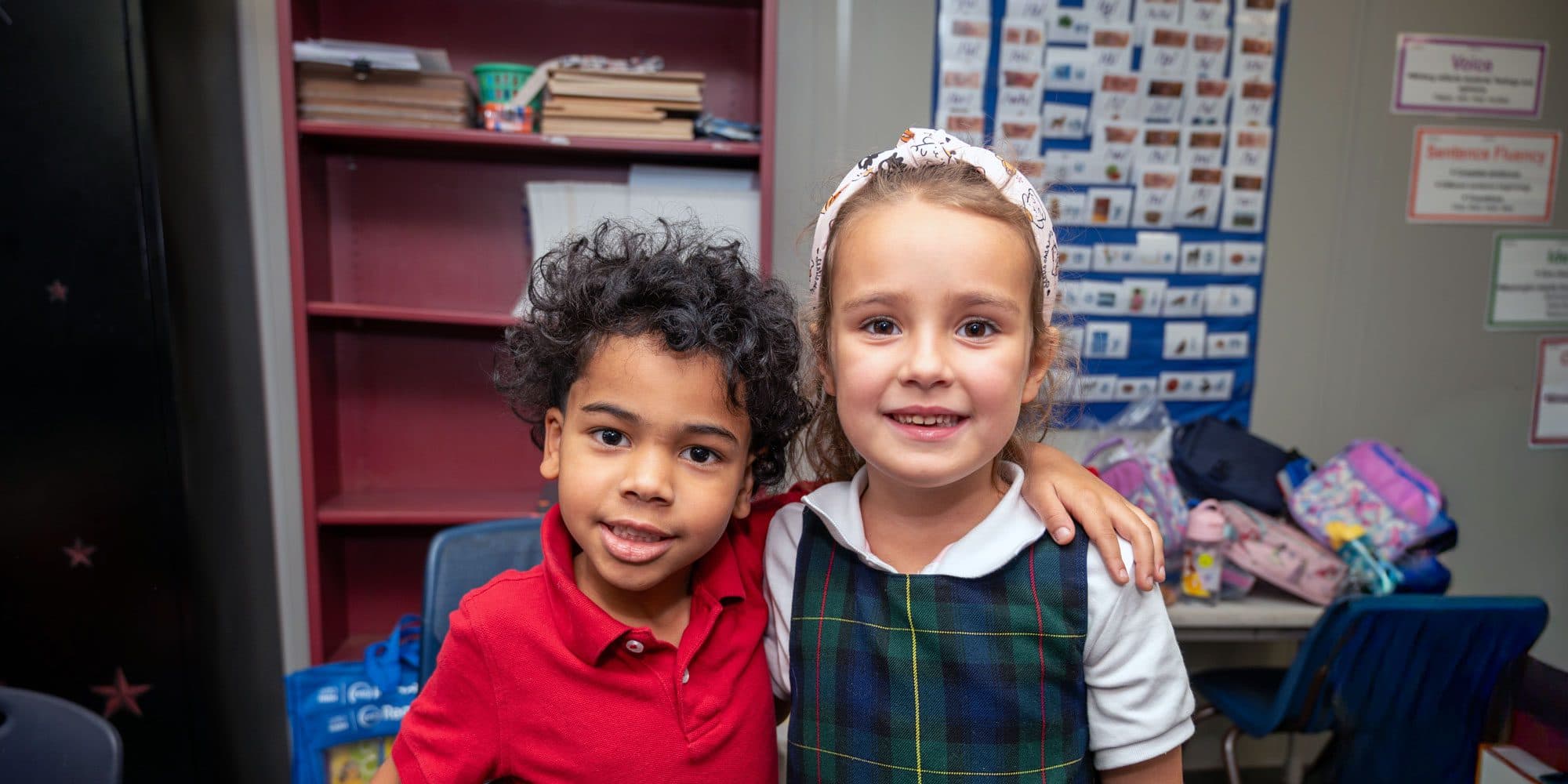 Two young students stand side by side and smile in a classroom.