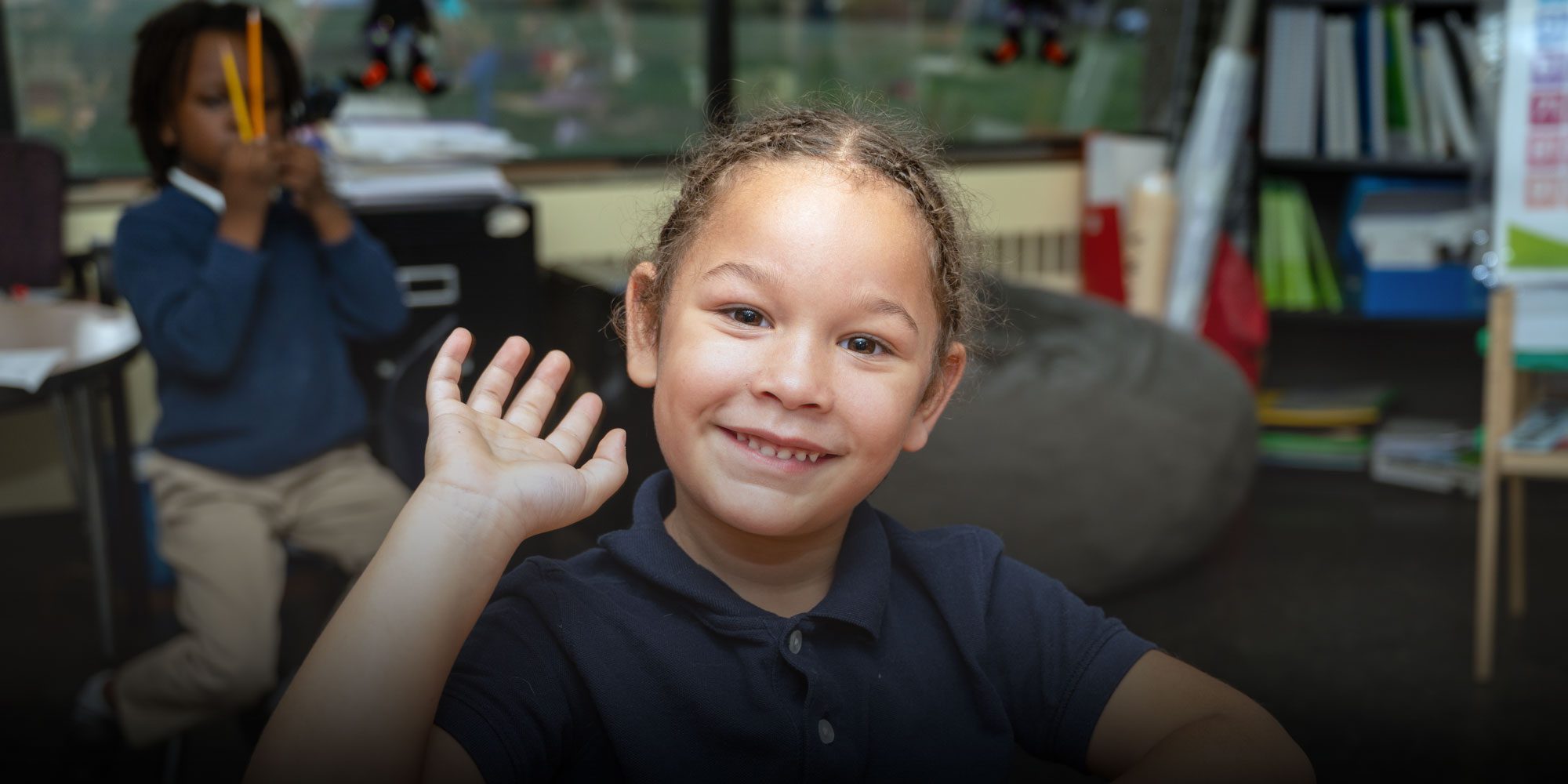 Smiling student in a navy school polo waves in a classroom.