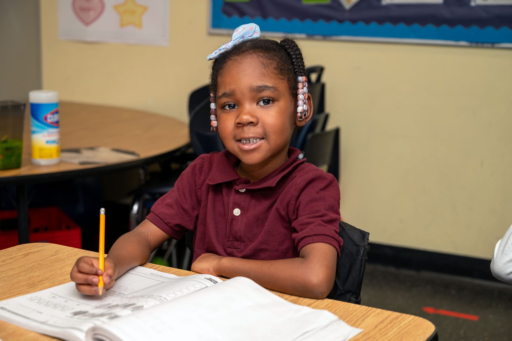 Young student holds a pencil over a workbook at a classroom desk.
